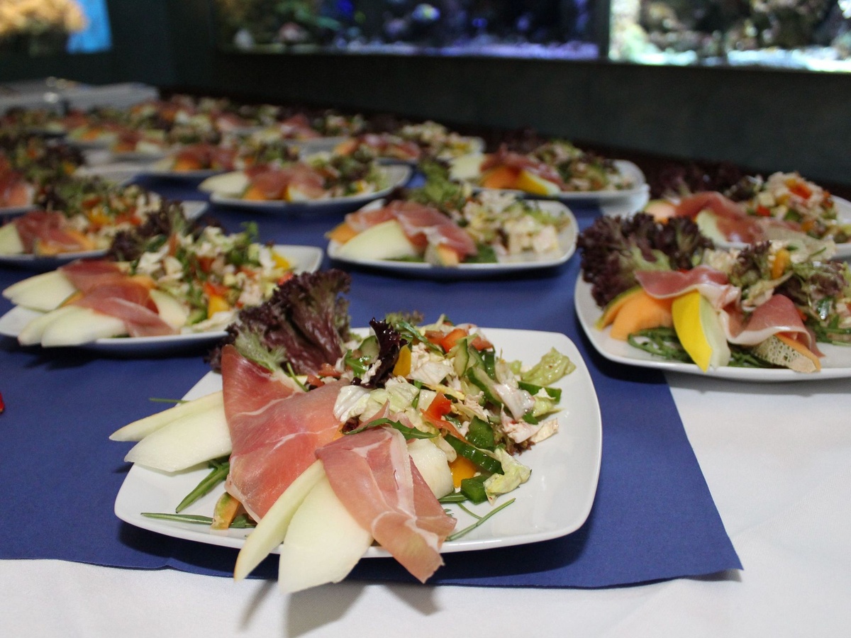 Mehrere Teller mit Schinken, Melone und Salat auf einer blau-weißen Tischdecke.