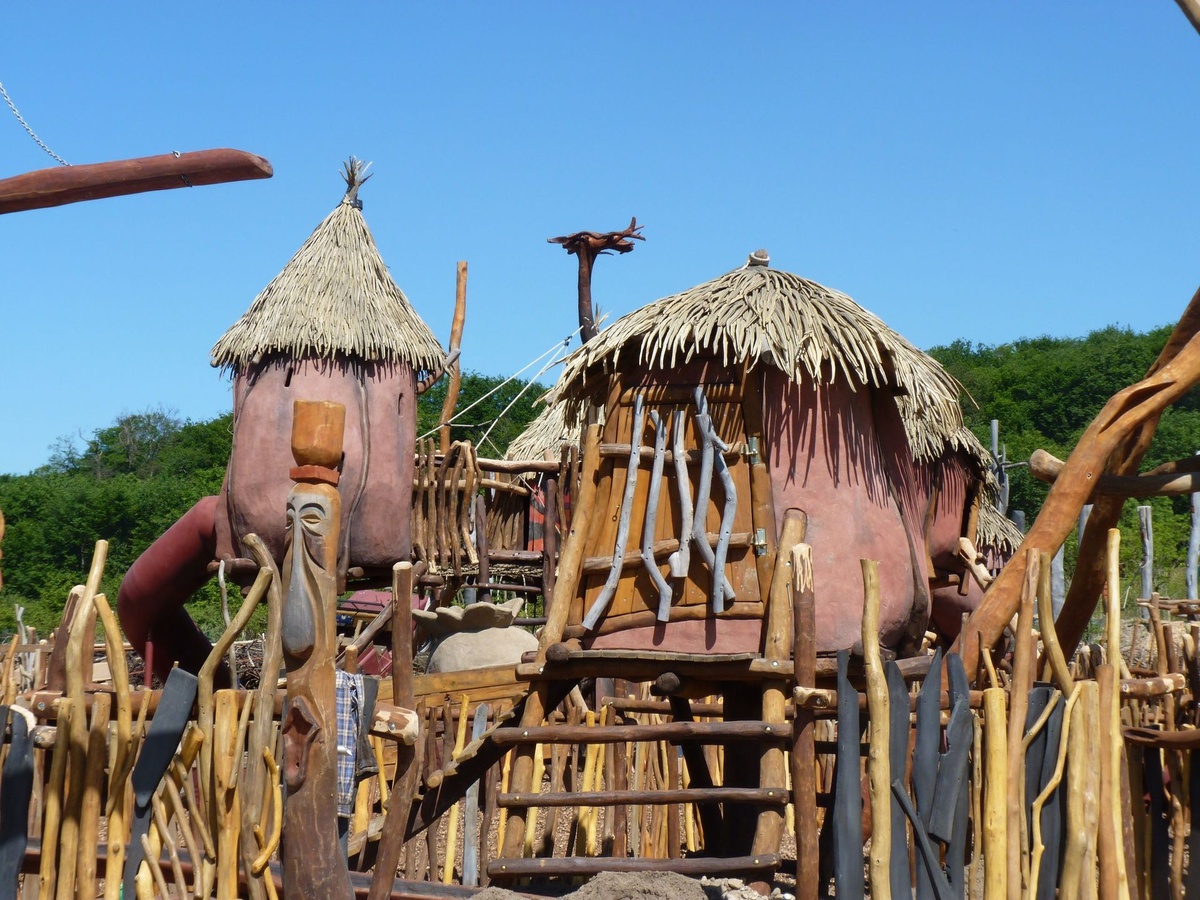 Spielplatz mit Strohdach-Hütten, Holztreppen und geschnitztem Totempfahl vor blauem Himmel.
