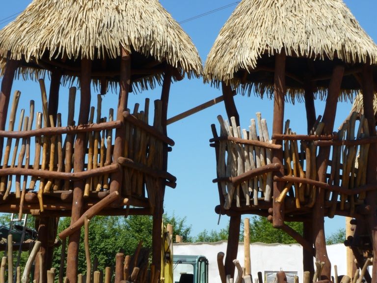 Zwei Holztürme mit Strohdächern, Geländern aus Rundholz, vor blauem Himmel.