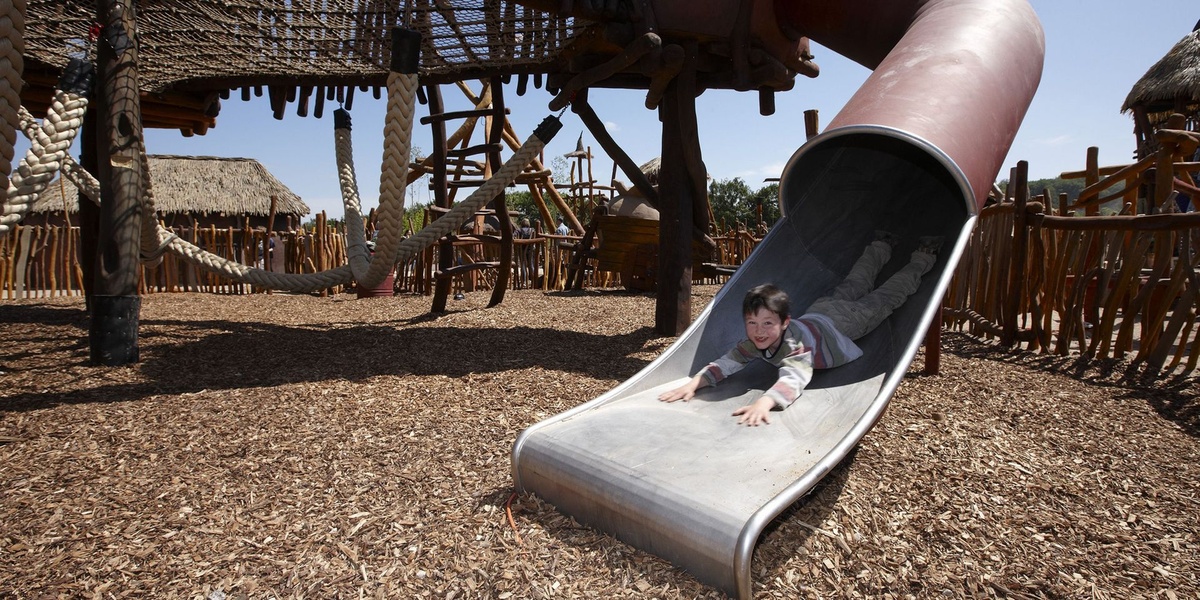 Kind rutscht lachend auf einer Rutsche im Holz-Spielplatz mit Seilnetzen und Strohdachhütten.