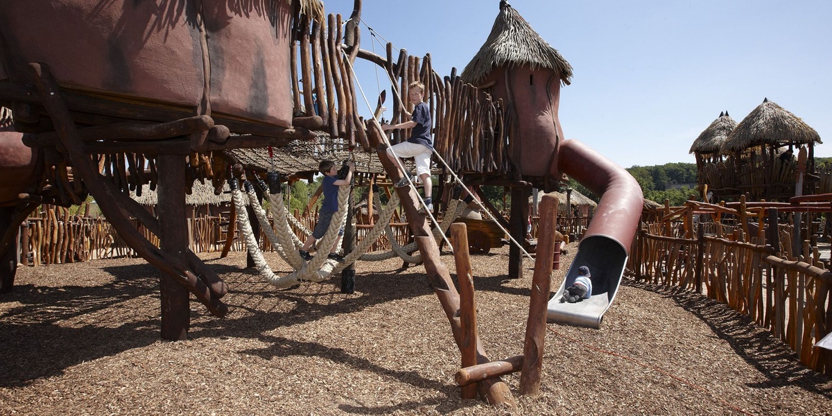 Kinder spielen auf Stelzenhütten mit Strohdächern, Rutsche und Kletterseilen.