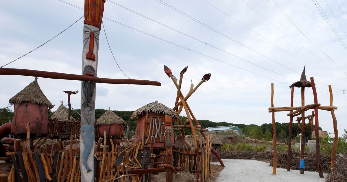 Spielplatz- oder Freizeitparkbereich mit Holzstangen-Zaun, Totem-Pfahl und Strohdach-Hütten unter bewölktem Himmel.