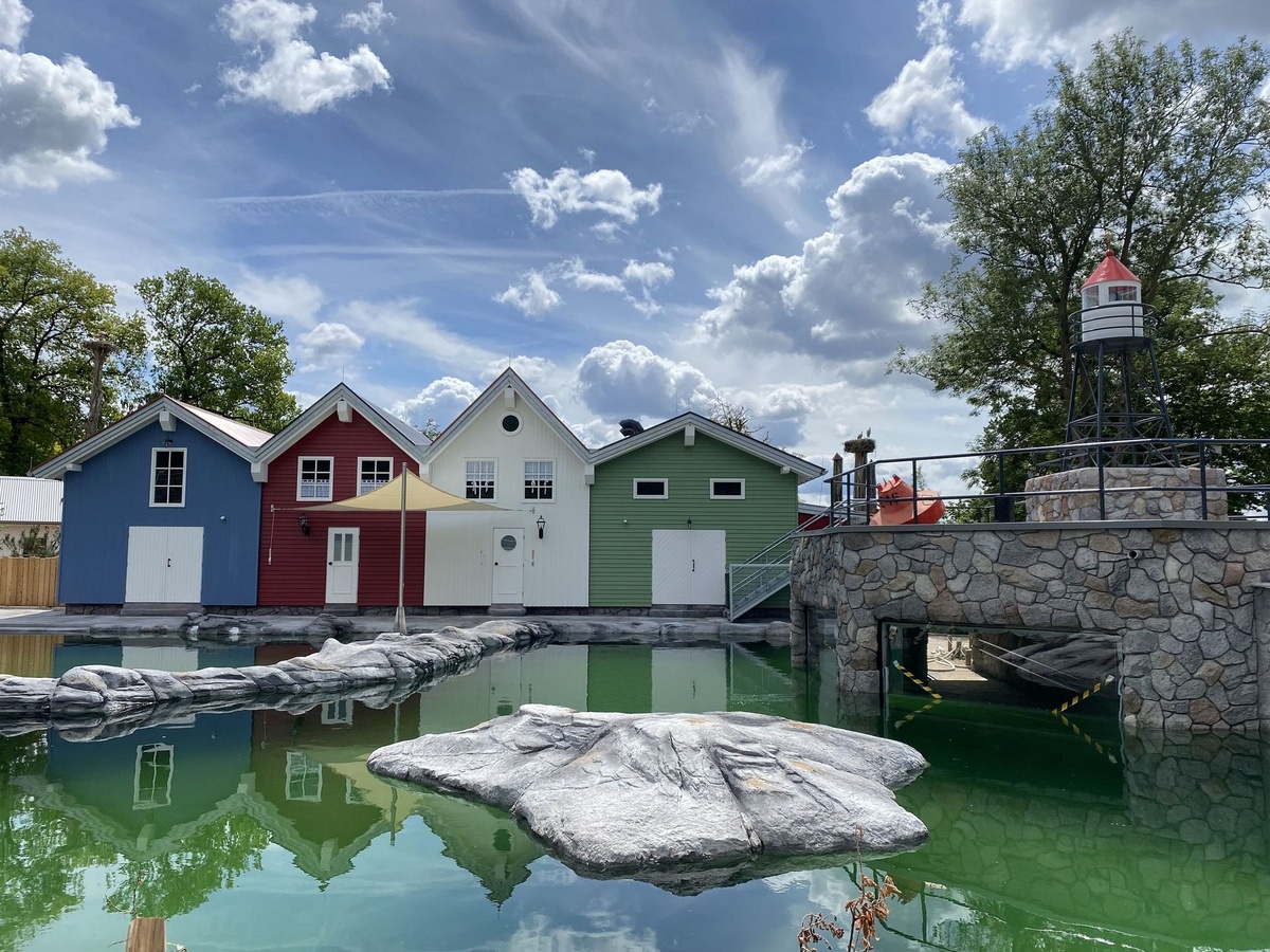 Drei bunte Häuser am Wasser. Davor Felsen und ein kleiner Leuchtturm auf Stein.