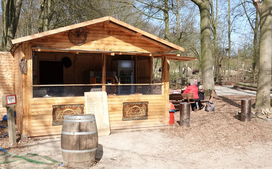 Kleine Holzhütte mit Theke und Menütafel, davor Holzfass und sitzende Personen auf Bank.