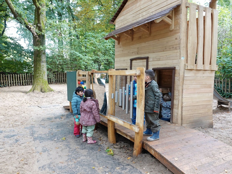 Holzspielhaus mit Rampe im Sand, mehrere Kinder spielen draußen und drinnen.