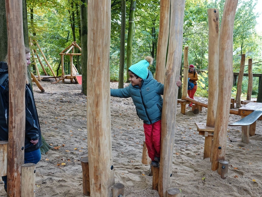 Kind balanciert auf Holzstämmen im Sandspielplatz. Die Frau schaut aufmerksam zu.