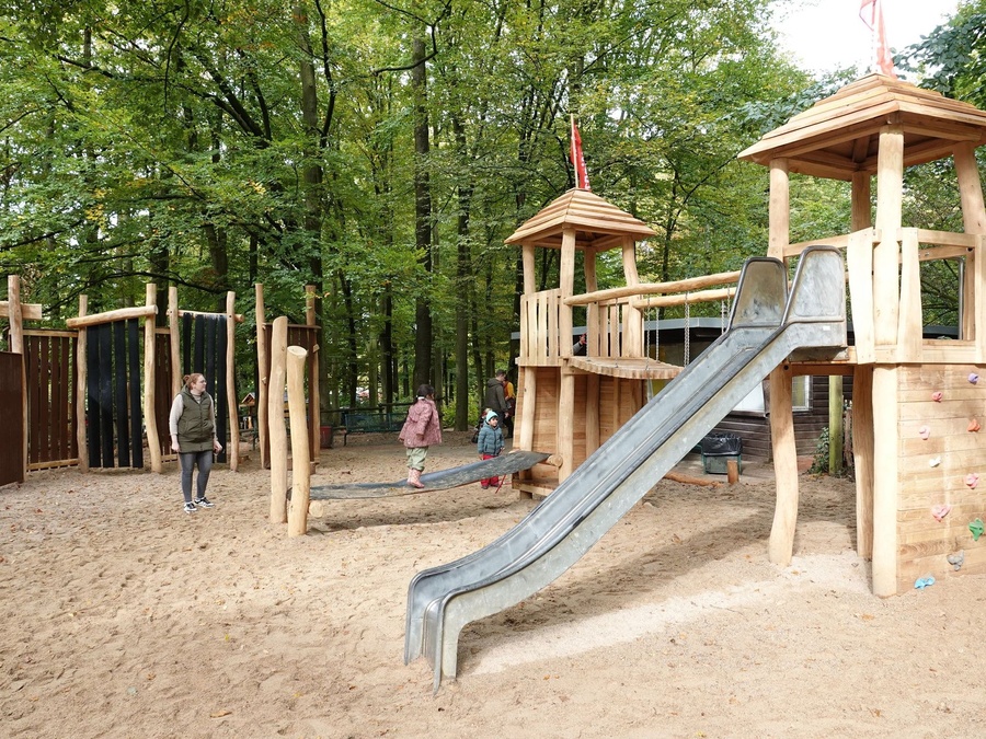 Kinder spielen auf Sandspielplatz mit Holzgerüst, Rutsche und Hangelbrücke im Wald.