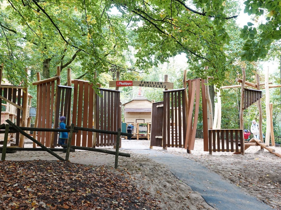 Ein Spielplatz im Wald mit Holzgerüst und Sandfläche. Kinder und Erwachsene spielen.