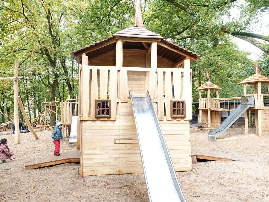 Holzspielplatz im Wald mit Rutsche, Türmen und Kindern, Sand bedeckt den Boden.