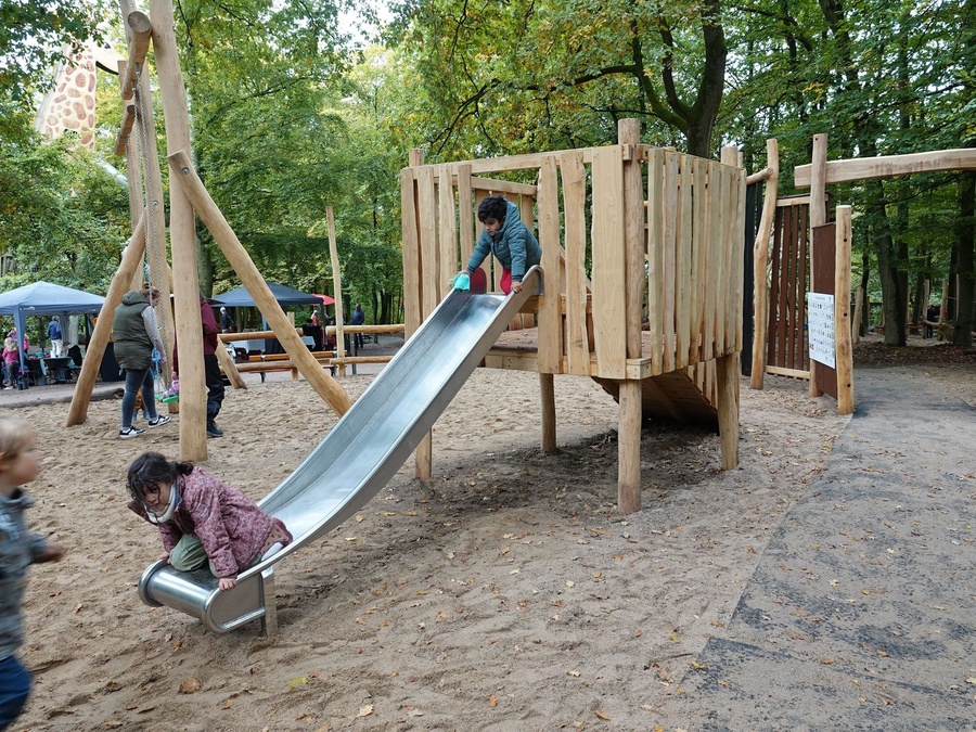 Kinder spielen auf einer Holzrutsche im Sand. Im Hintergrund stehen Bäume und Besucher.