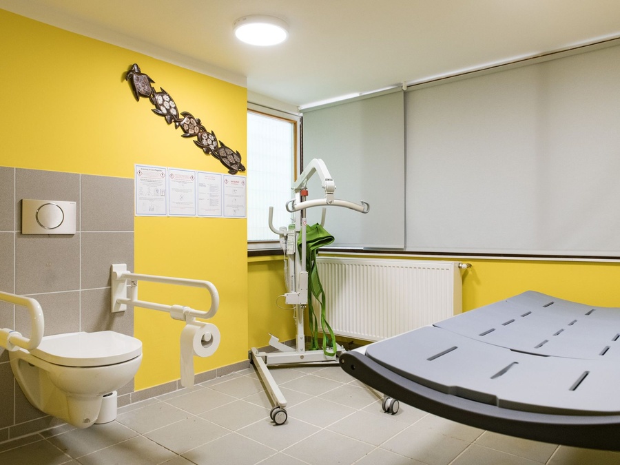 Barrierefreies WC mit Haltegriffen, Pflegeliege, Patientenlifter und eine gelbe Wand.
