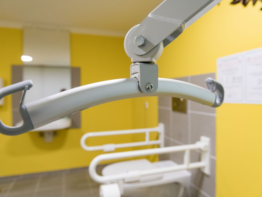 Barrierefreies Bad: Nahaufnahme eines Lifters vor gelber Wand, Toilette mit Haltegriffe.