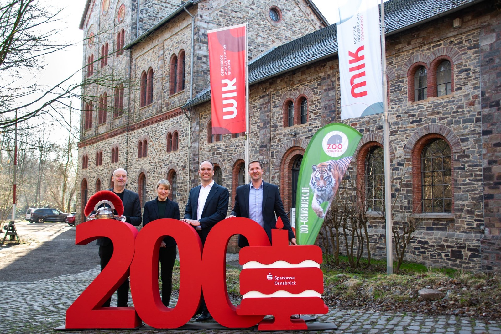 Vier Personen vor roter 200. Im Hintergrund sind drei Fahnen und ein Gebäude.